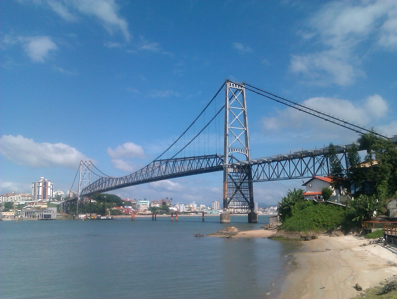 Ponte Hercílio Luz em Florianópolis, Santa Catarina, com prédios ao fundo, faixa de areia à direita e céu azul